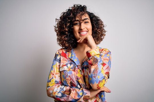 Young beautiful curly arab woman wearing floral colorful shirt standing over white background looking confident at the camera with smile with crossed arms and hand raised on chin. Thinking positive.