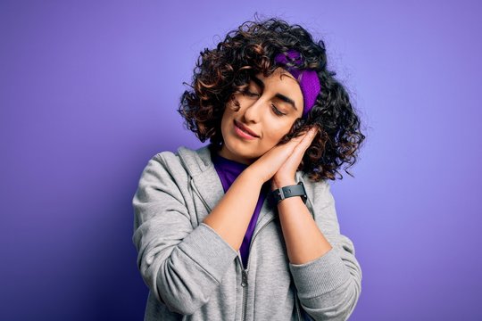 Beautiful curly arab sportswoman doing sport wearing sportswear over purple background sleeping tired dreaming and posing with hands together while smiling with closed eyes.