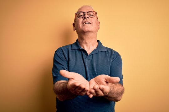 Middle age handsome hoary man wearing casual polo and glasses over yellow background Smiling with hands palms together receiving or giving gesture. Hold and protection