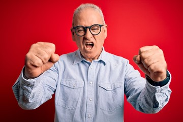 Middle age handsome hoary man wearing casual striped shirt and glasses over red background angry and mad raising fists frustrated and furious while shouting with anger. Rage and aggressive concept.