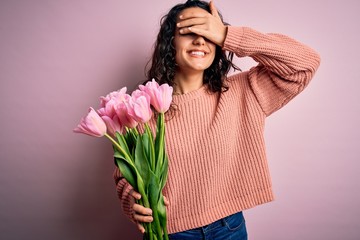 Young beautiful romantic woman with curly hair holding bouquet of pink tulips smiling and laughing with hand on face covering eyes for surprise. Blind concept.