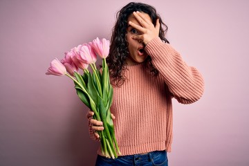 Young beautiful romantic woman with curly hair holding bouquet of pink tulips peeking in shock covering face and eyes with hand, looking through fingers with embarrassed expression.