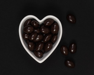 Dark chocolate candy covered almonds in a heart shaped bowl on a black background.