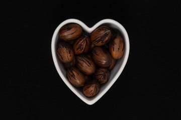 Pecan nuts with the nutshell in a heart shaped bowl on a black background.