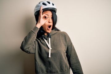 Young beautiful chinese woman wearing bike helmet over isolated white background doing ok gesture shocked with surprised face, eye looking through fingers. Unbelieving expression.