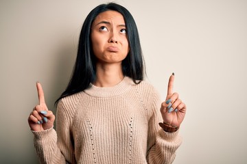 Young beautiful chinese woman wearing casual sweater over isolated white background Pointing up looking sad and upset, indicating direction with fingers, unhappy and depressed.