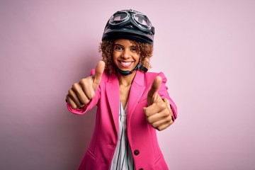 African american motorcyclist woman with curly hair wearing moto helmet over pink background approving doing positive gesture with hand, thumbs up smiling and happy for success. Winner gesture.