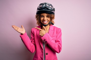 African american motorcyclist woman with curly hair wearing moto helmet over pink background amazed and smiling to the camera while presenting with hand and pointing with finger.