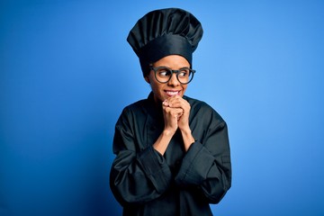 Young african american chef woman wearing cooker uniform and hat over blue background laughing nervous and excited with hands on chin looking to the side