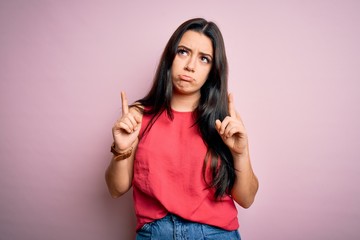 Young brunette woman wearing casual summer shirt over pink isolated background Pointing up looking sad and upset, indicating direction with fingers, unhappy and depressed.