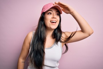 Young brunette woman wearing casual sport cap over pink background very happy and smiling looking far away with hand over head. Searching concept.