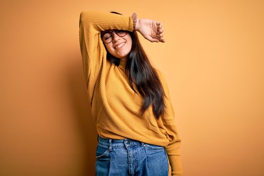 Young brunette woman wearing glasses and casual sweater over yellow isolated background covering eyes with arm smiling cheerful and funny. Blind concept.