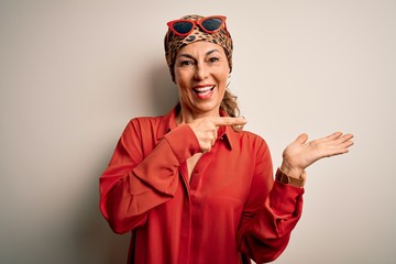 Middle age brunette woman wearing handkerchief on head and shirt over white background amazed and smiling to the camera while presenting with hand and pointing with finger.