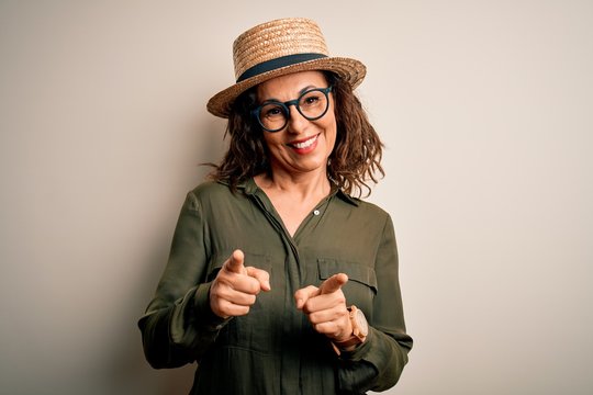 Middle age brunette woman wearing glasses and hat standing over isolated white background pointing fingers to camera with happy and funny face. Good energy and vibes.