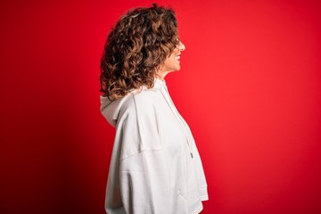 Middle age beautiful sporty woman wearing white sweatshirt over isolated red background looking to side, relax profile pose with natural face with confident smile.