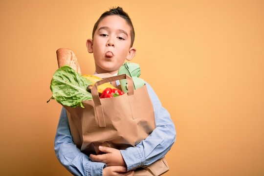 Adorable toddler holding paper bag with food standing over isolated yellow background