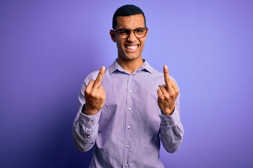 Fototapeta premium Handsome african american man wearing striped shirt and glasses over purple background Showing middle finger doing fuck you bad expression, provocation and rude attitude. Screaming excited