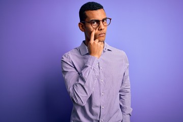 Obraz premium Handsome african american man wearing striped shirt and glasses over purple background Pointing to the eye watching you gesture, suspicious expression