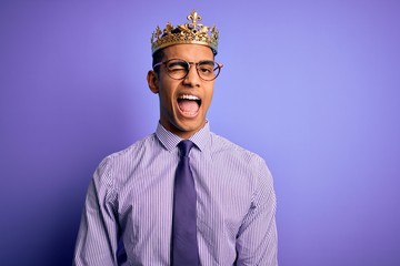 Young handsome african american man wearing golden crown of king over purple background winking looking at the camera with sexy expression, cheerful and happy face.