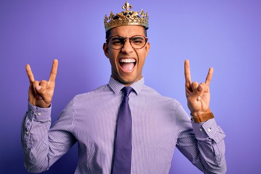 Young handsome african american man wearing golden crown of king over purple background shouting with crazy expression doing rock symbol with hands up. Music star. Heavy concept.
