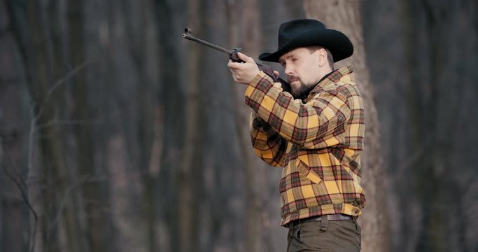 Male hunter in hat and black-yellow jacket shooting rifle in forest, hobby