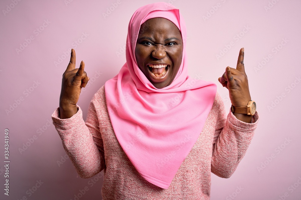 Sticker Young african american plus size woman wearing muslim hijab over isolated pink background shouting with crazy expression doing rock symbol with hands up. Music star. Heavy concept.