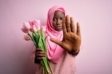 Young african american plus size woman wearing muslim hijab holding bouquet of pink tulips doing stop sing with palm of the hand. Warning expression with negative and serious gesture on the face.