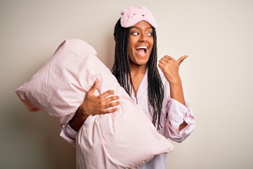 Young african american woman wearing pajama and eye mask hugging bed pillow pointing and showing...