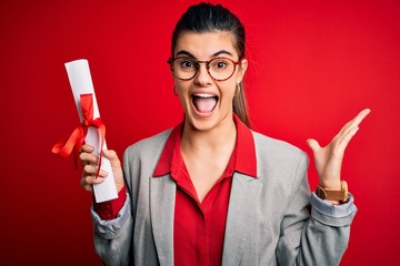 Young beautiful brunette student woman wearing glasses holding university degree diploma very happy and excited, winner expression celebrating victory screaming with big smile and raised hands