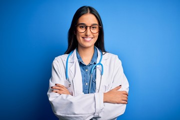 Young beautiful doctor woman wearing stethoscope and glasses over blue background happy face smiling with crossed arms looking at the camera. Positive person.