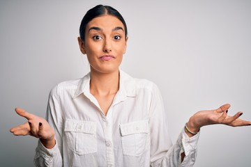 Young beautiful brunette woman wearing casual shirt over isolated white background clueless and...