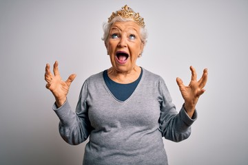 Senior beautiful grey-haired woman wearing golden queen crown over white background crazy and mad shouting and yelling with aggressive expression and arms raised. Frustration concept.