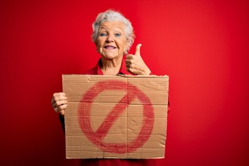 Senior beautiful grey-haired woman holding banner with prohibited signal over red background happy with big smile doing ok sign, thumb up with fingers, excellent sign