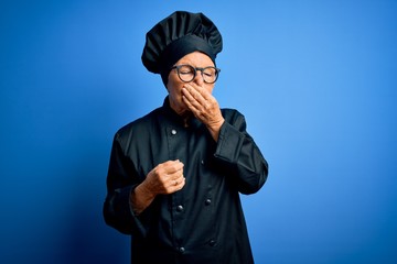 Senior beautiful grey-haired chef woman wearing cooker uniform and hat over blue background smelling something stinky and disgusting, intolerable smell, holding breath with fingers on nose. Bad smell