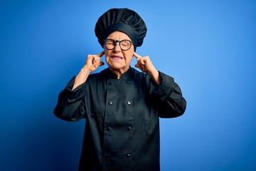 Senior beautiful grey-haired chef woman wearing cooker uniform and hat over blue background covering ears with fingers with annoyed expression for the noise of loud music. Deaf concept.