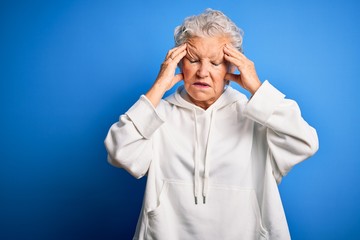 Senior beautiful sporty woman wearing white sweatshirt over isolated blue background suffering from headache desperate and stressed because pain and migraine. Hands on head.