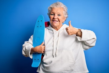 Senior beautiful sporty woman holding skate standing over isolated blue background happy with big smile doing ok sign, thumb up with fingers, excellent sign © Krakenimages.com
