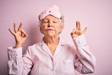 Senior beautiful woman wearing sleep mask and pajama over isolated pink background relax and smiling with eyes closed doing meditation gesture with fingers. Yoga concept.