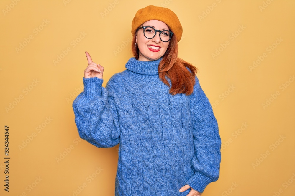 Poster Young beautiful redhead woman wearing french beret and glasses over yellow background with a big smile on face, pointing with hand and finger to the side looking at the camera.