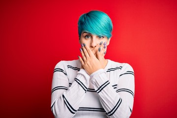 Young beautiful woman with blue fashion hair wearing striped sweater over red background shocked covering mouth with hands for mistake. Secret concept.