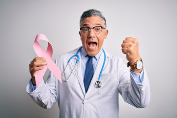 Middle age handsome grey-haired doctor man holding pink cancer ribbon screaming proud and celebrating victory and success very excited, cheering emotion