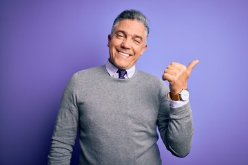 Middle age handsome grey-haired man wearing elegant sweater over purple background smiling with happy face looking and pointing to the side with thumb up.