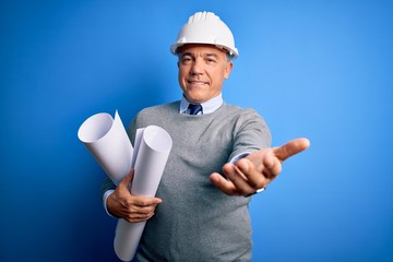 Middle age handsome grey-haired architect man wearing safety helmet holding blueprints smiling cheerful offering palm hand giving assistance and acceptance.