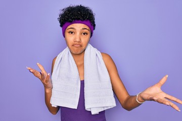 Beautiful african american sporty woman doing sport wearing towel over purple background clueless and confused expression with arms and hands raised. Doubt concept.