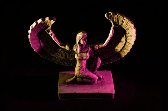 Closeup to a Isis. female old winged egyptian god mini figurine iluminated with yellow and pink lights over black background 