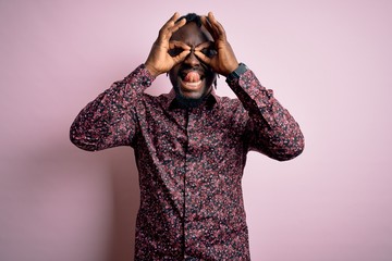 Young handsome african american man wearing casual shirt standing over pink background doing ok gesture like binoculars sticking tongue out, eyes looking through fingers. Crazy expression.