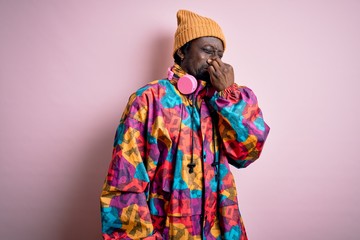 Young handsome african american man wearing colorful coat and cap over pink background smelling something stinky and disgusting, intolerable smell, holding breath with fingers on nose. Bad smell