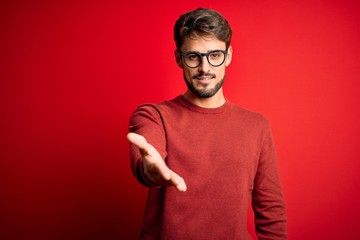 Young handsome man with beard wearing glasses and sweater standing over red background smiling...
