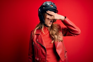 Young beautiful brunette motrocyclist woman wearing moto helmet over red background peeking in shock covering face and eyes with hand, looking through fingers with embarrassed expression.