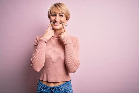 Young blonde woman with short hair wearing casual turtleneck sweater over pink background Smiling with open mouth, fingers pointing and forcing cheerful smile